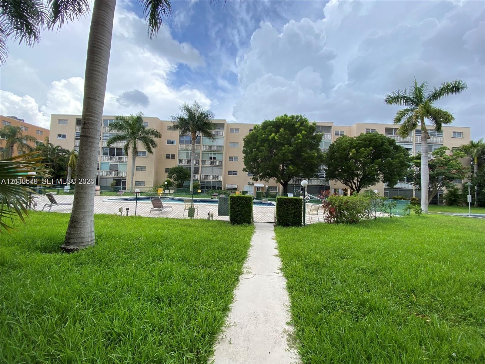 Ground-level recreational area featuring a paved walkway, swimming pool, manicured lawn, mature palm trees, and professional landscaping