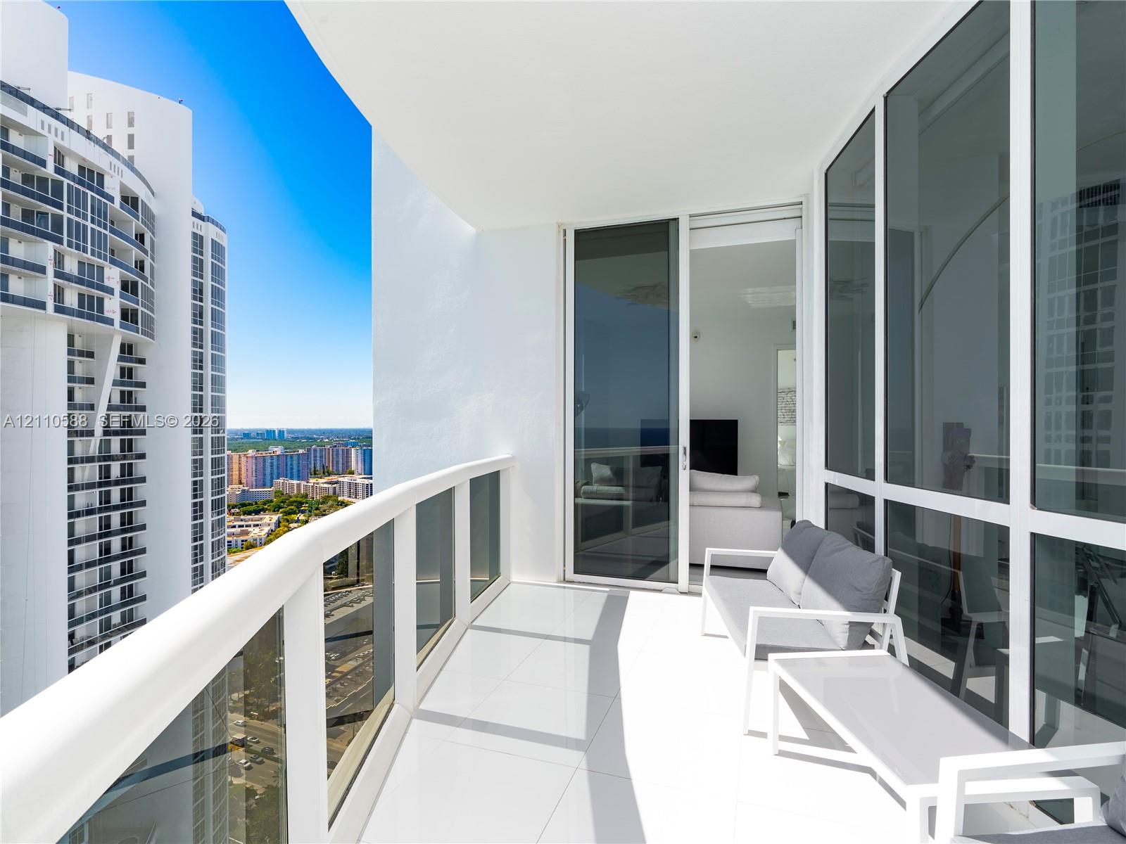 Covered balcony with white tile flooring and glass panel railing, featuring floor-to-ceiling glass doors