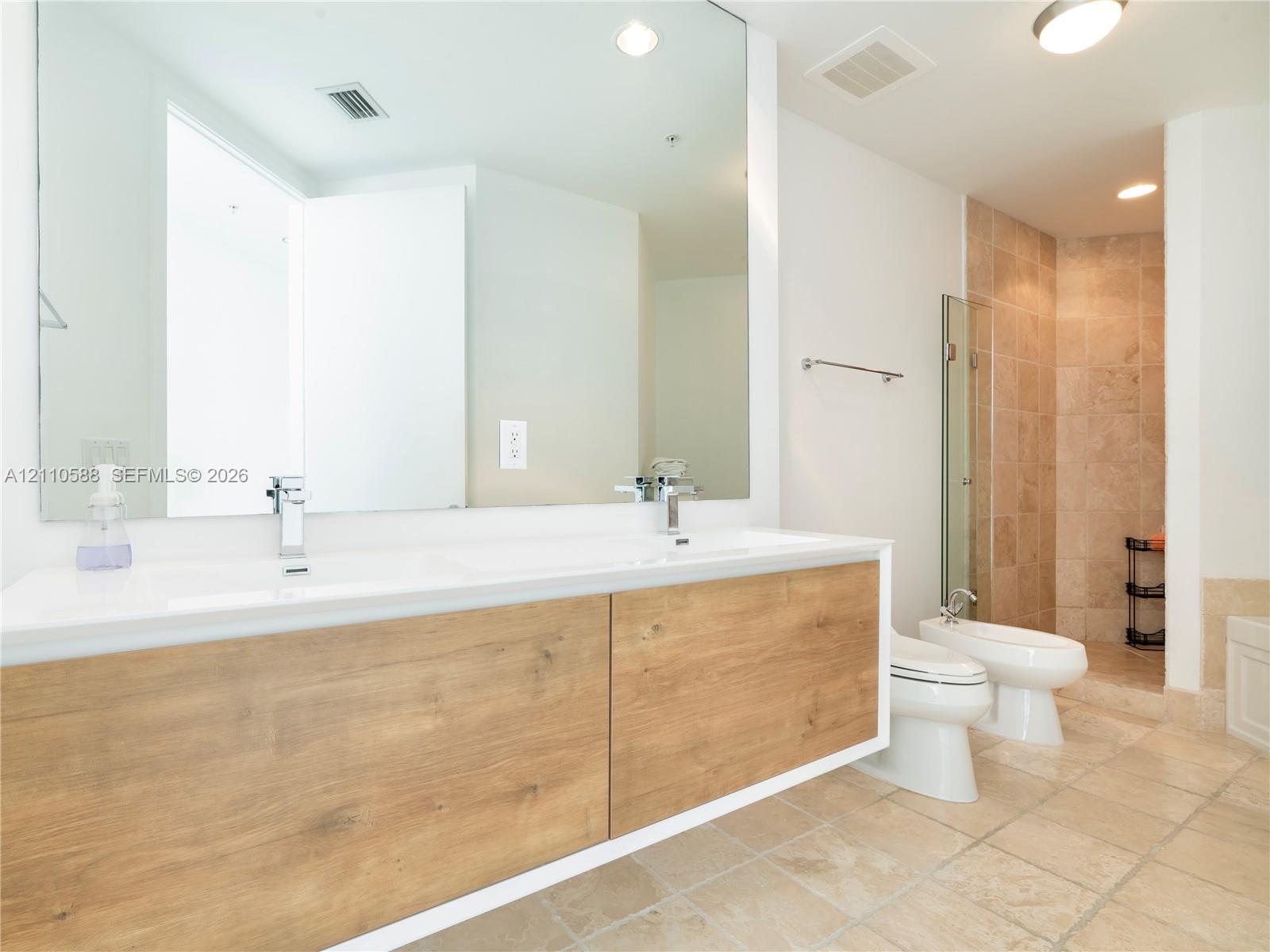 Floating dual-sink vanity with wood-finish cabinetry, expansive wall-mounted mirror, stone-tile flooring, and glass-enclosed shower