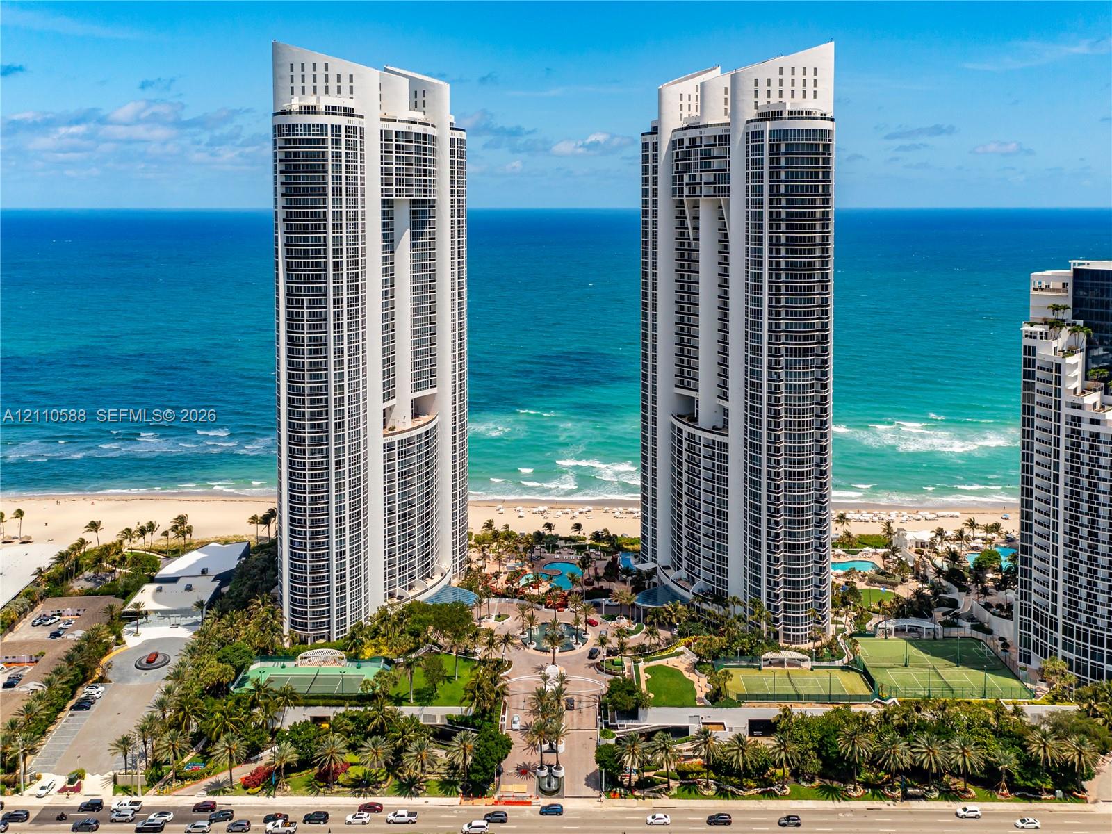 High-rise waterfront towers featuring expansive floor-to-ceiling windows, multiple hard-surface tennis courts, landscaped grounds with integrated pool areas, and direct sandy beach access