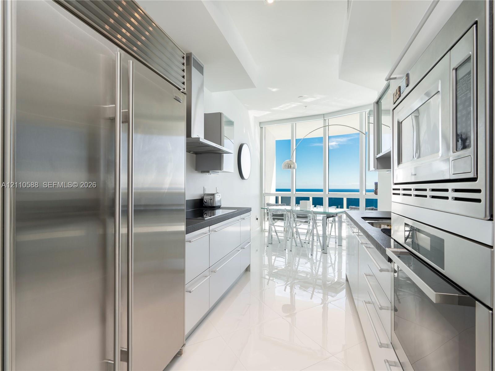 Modern galley kitchen featuring high-gloss cabinetry, stainless steel professional-grade appliances, dark stone countertops, polished tile flooring, and floor-to-ceiling windows