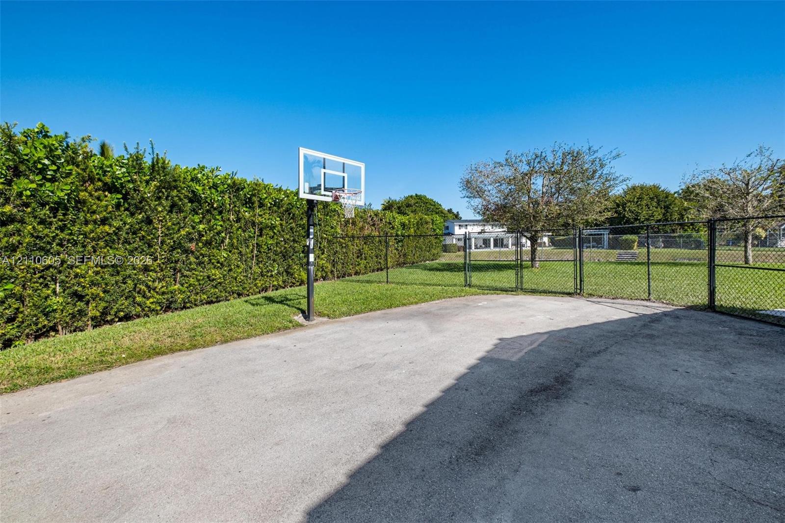 Paved recreational court featuring a built-in basketball hoop, enclosed by chain-link fencing and mature perimeter hedging
