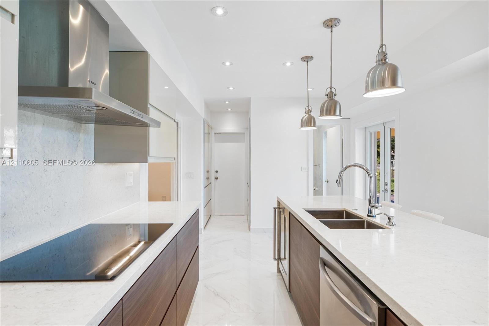 Modern kitchen featuring wood-finish flat-panel cabinetry, light-toned stone-look countertops, stainless steel appliances, marble-patterned flooring, and three pendant lights