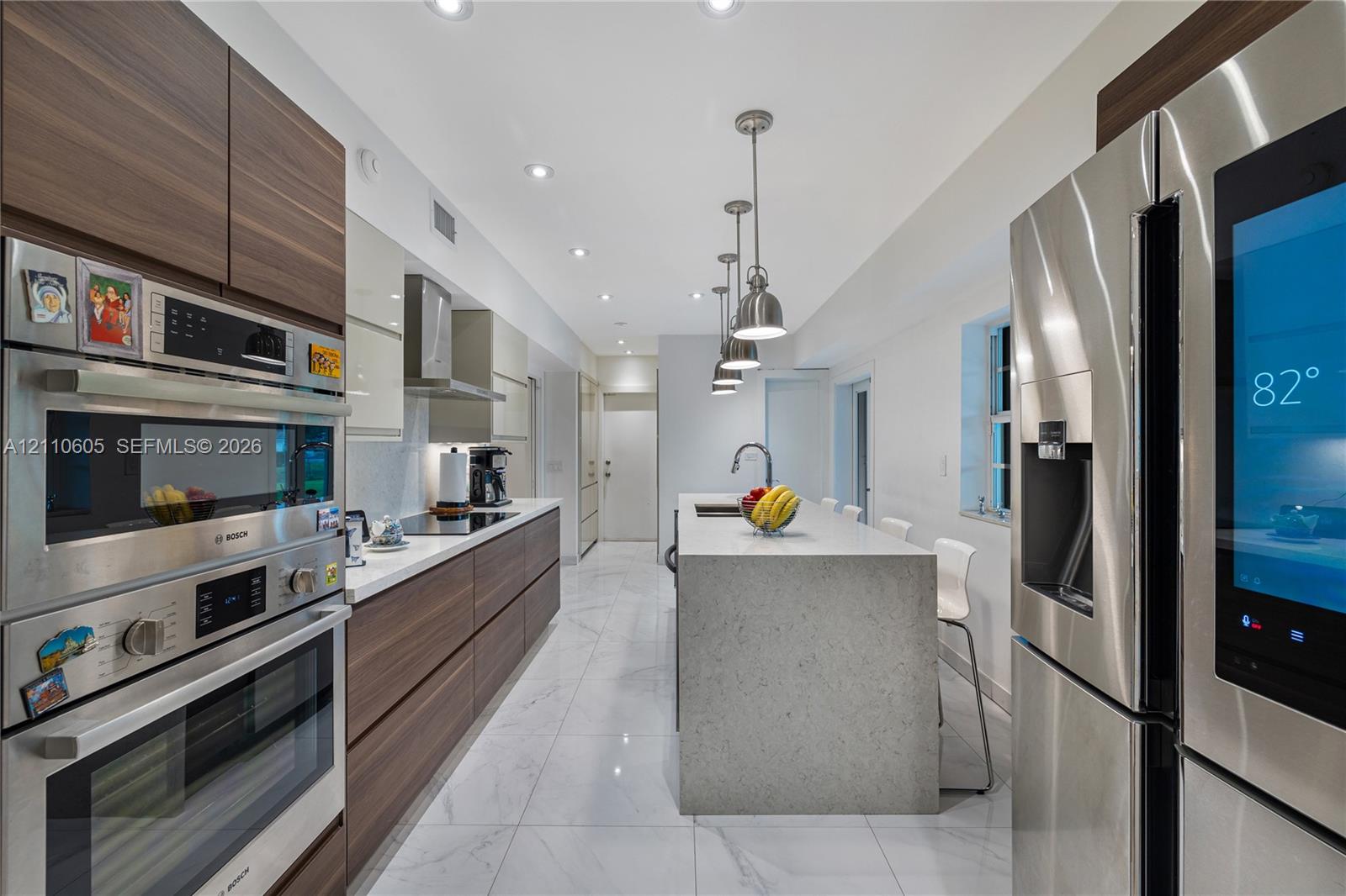 Modern kitchen featuring wood-finish cabinetry, a central island with a solid surface countertop, stainless steel appliances, large-format tile flooring, and pendant lighting