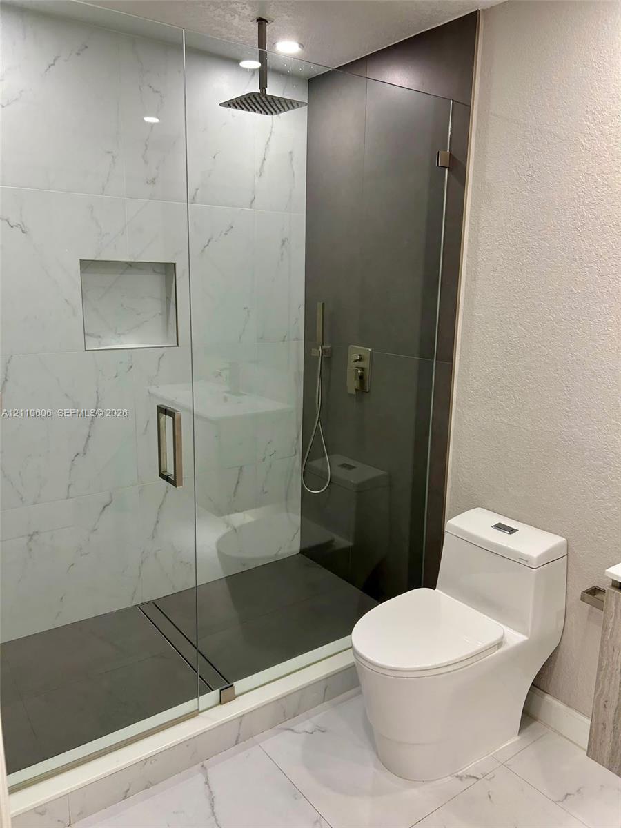 Walk-in shower featuring a frameless glass enclosure, rainfall showerhead, recessed storage niche, and contrast stone-finish tiling, paired with a modern dual-flush elongated toilet