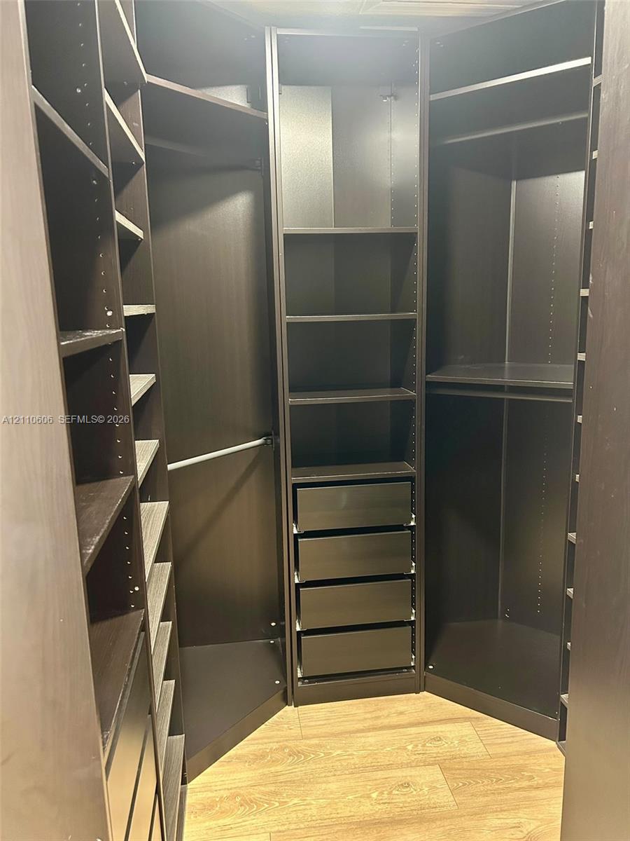 Custom walk-in closet featuring floor-to-ceiling dark cabinetry, adjustable shelving, integrated drawer stack, metal garment rods, and wood-finish flooring