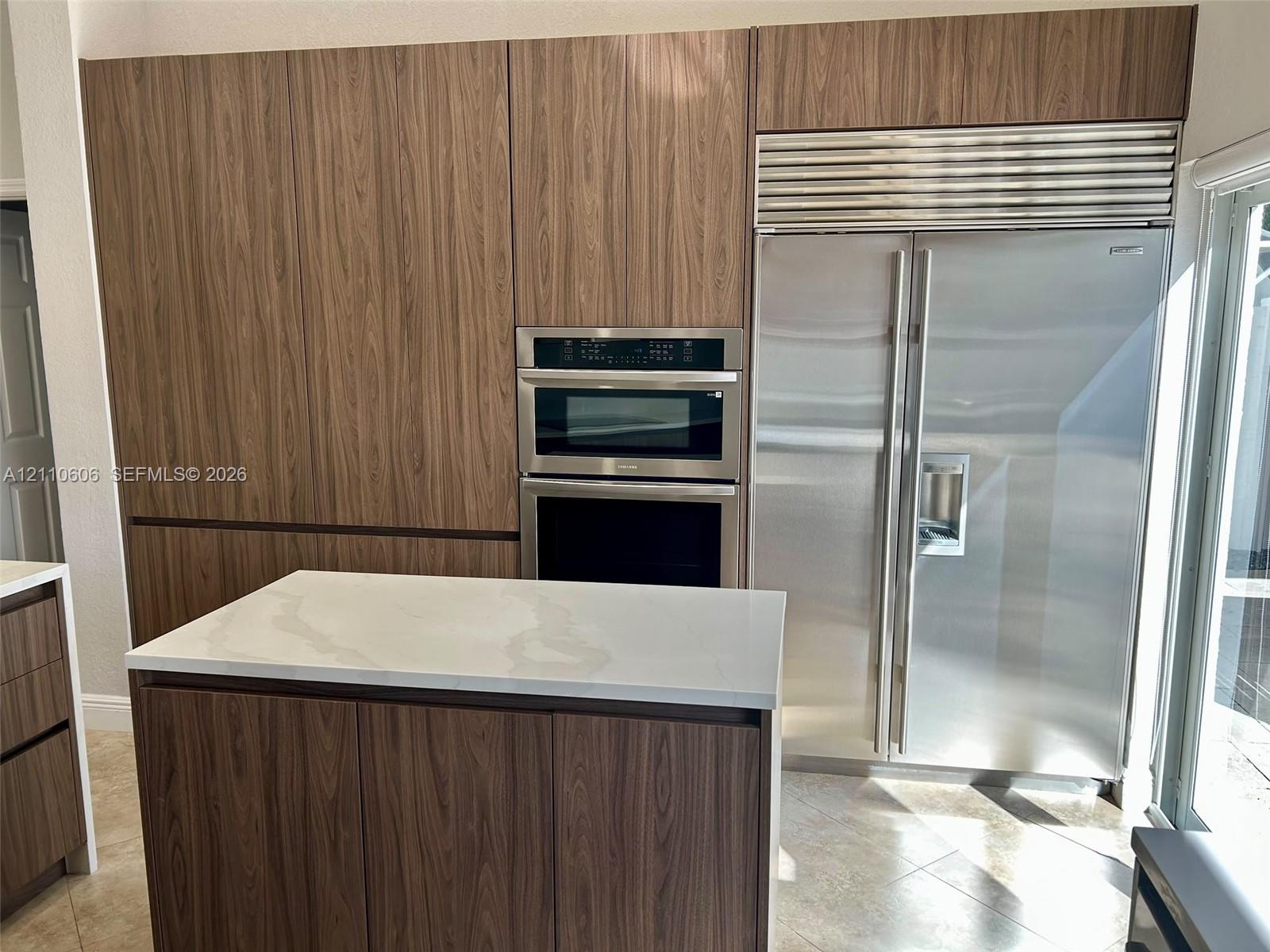 Kitchen featuring floor-to-ceiling wood-finish cabinetry, a built-in stainless steel refrigerator, a wall-mounted microwave and oven combination, and a central island topped with a white stone-surface countertop
