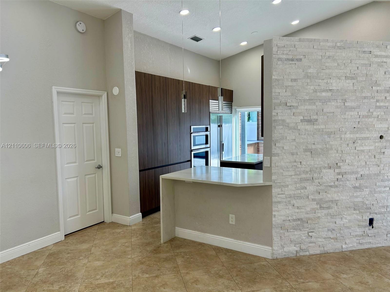 Kitchen featuring flat-panel wood-finish cabinetry, built-in double ovens, a peninsula with a waterfall-edge countertop, and a decorative stone-veneer accent wall