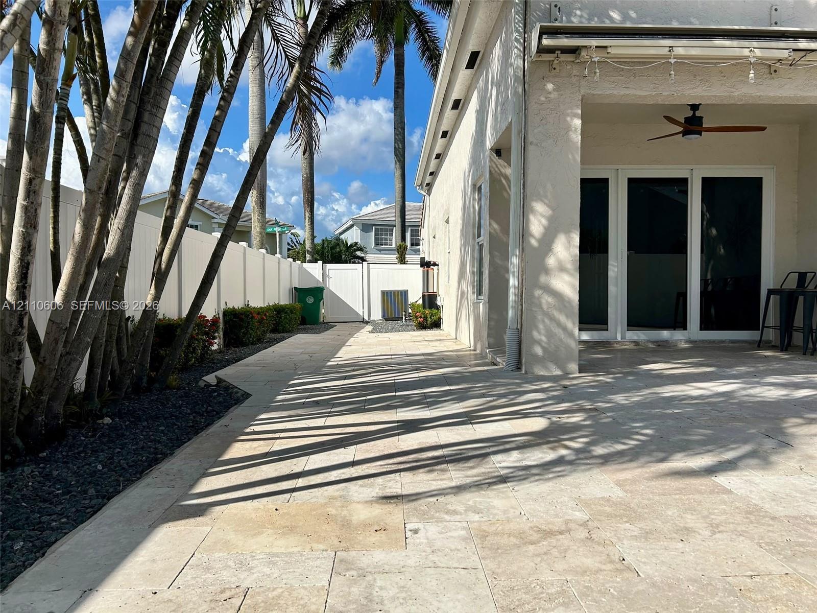 Covered patio featuring stone-tiled flooring and a ceiling fan, complemented by a white vinyl privacy fence and mature palm trees