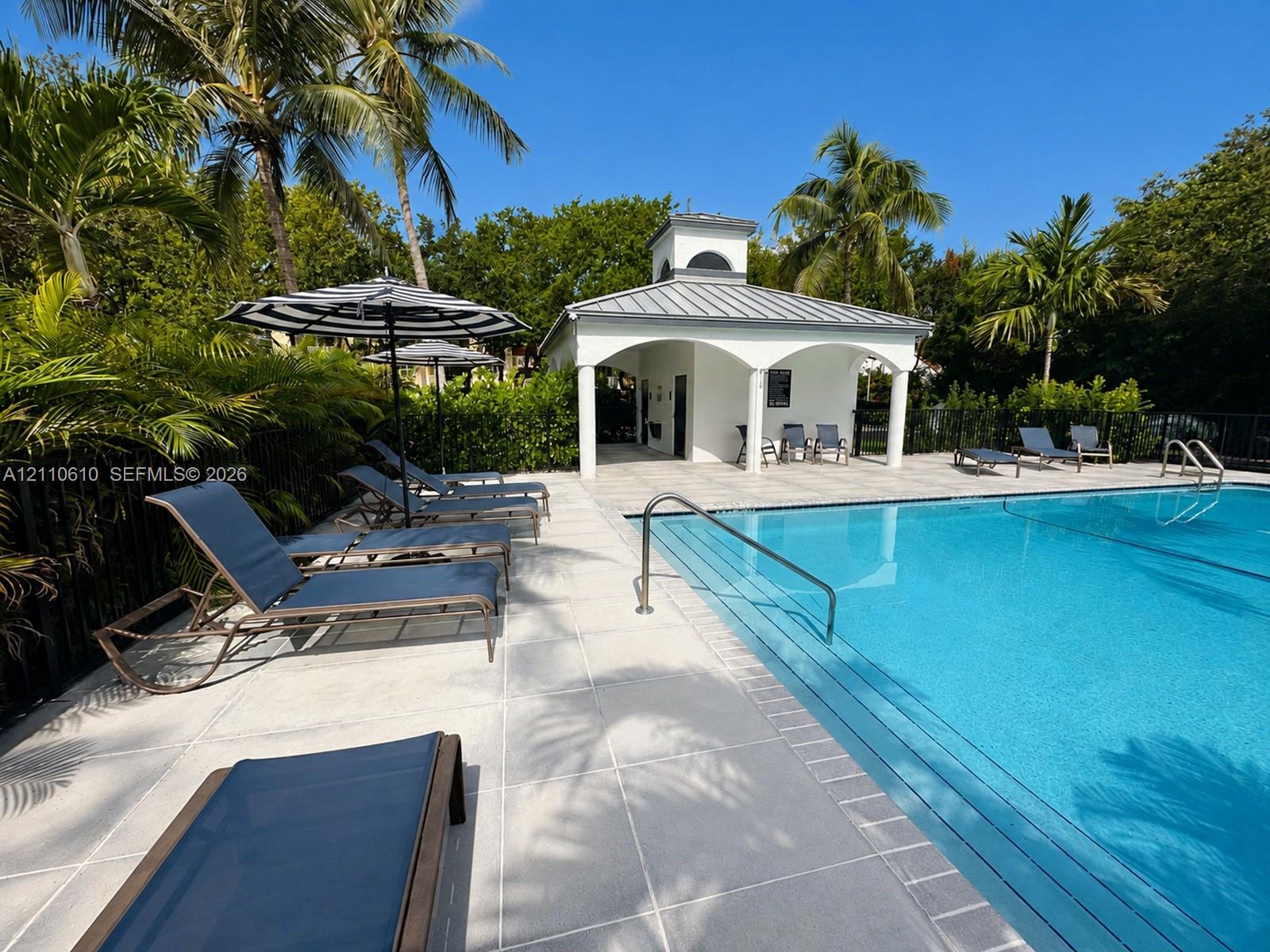 In-ground swimming pool with stone-tile decking, covered pavilion with arched supports, metal roof, and metal perimeter fencing