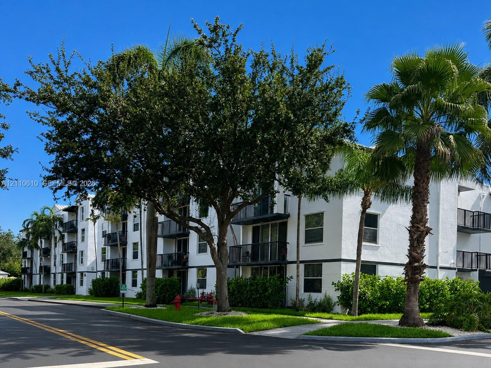 Multi-story exterior featuring white stucco finish, dark-toned balconies, and mature palm and shade trees set within a landscaped lawn