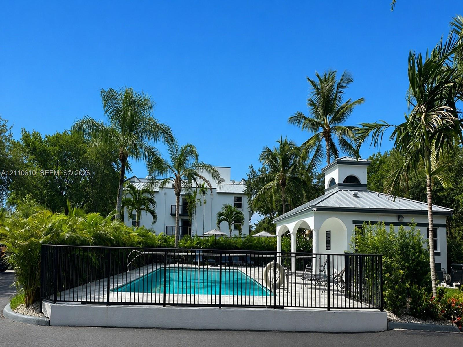 Swimming pool enclosed by black metal fencing, featuring a detached cabana with arched architectural details and a metal roof, surrounded by mature palm trees and manicured shrubbery