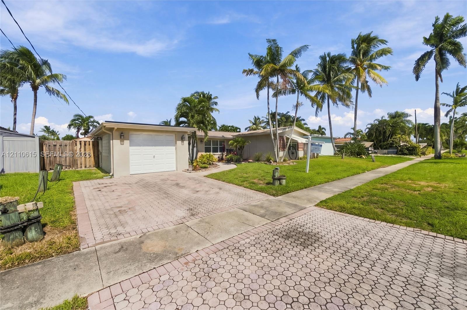 Single-story exterior featuring a paved driveway, one-car garage, manicured lawn, and mature palm trees