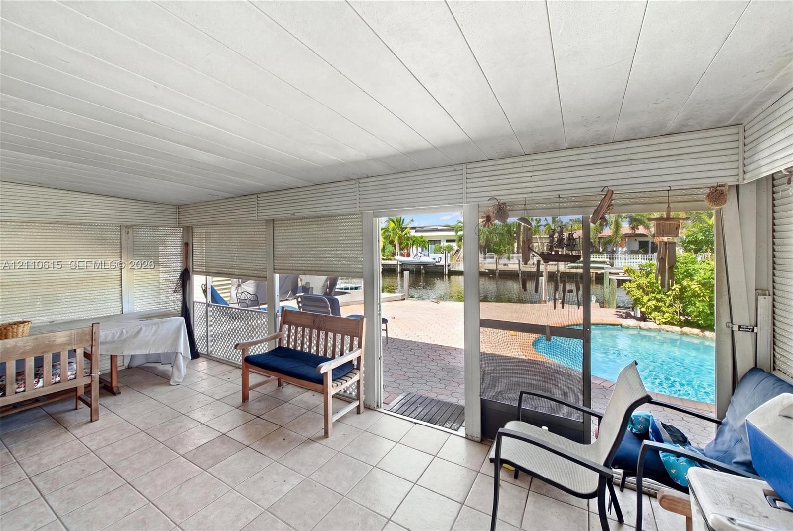 Enclosed patio featuring tile flooring, horizontal slat wall finishes, ceiling panels, and direct access to a paved pool deck with a waterfront bulkhead