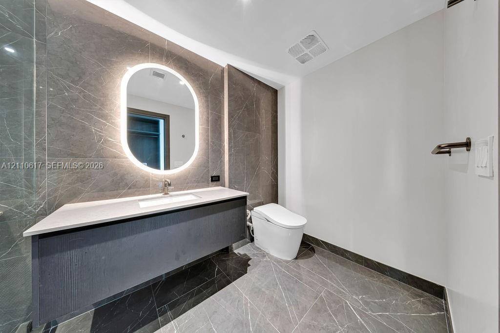 Floating vanity featuring a stone-topped counter and integrated sink, illuminated oval wall mirror, large-format stone tile wall and flooring, and wall-mounted toilet