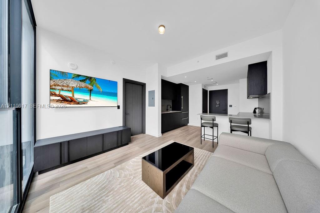 Open-concept living space featuring wood-finish flooring, floor-to-ceiling windows, modern matte black cabinetry, and a kitchen island with a stone-finish countertop