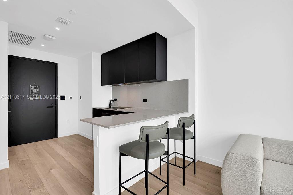 Kitchen area featuring matte black cabinetry, light-toned stone-finish countertops, a matching full-height backsplash, wood-finish flooring, and a black solid-core entry door