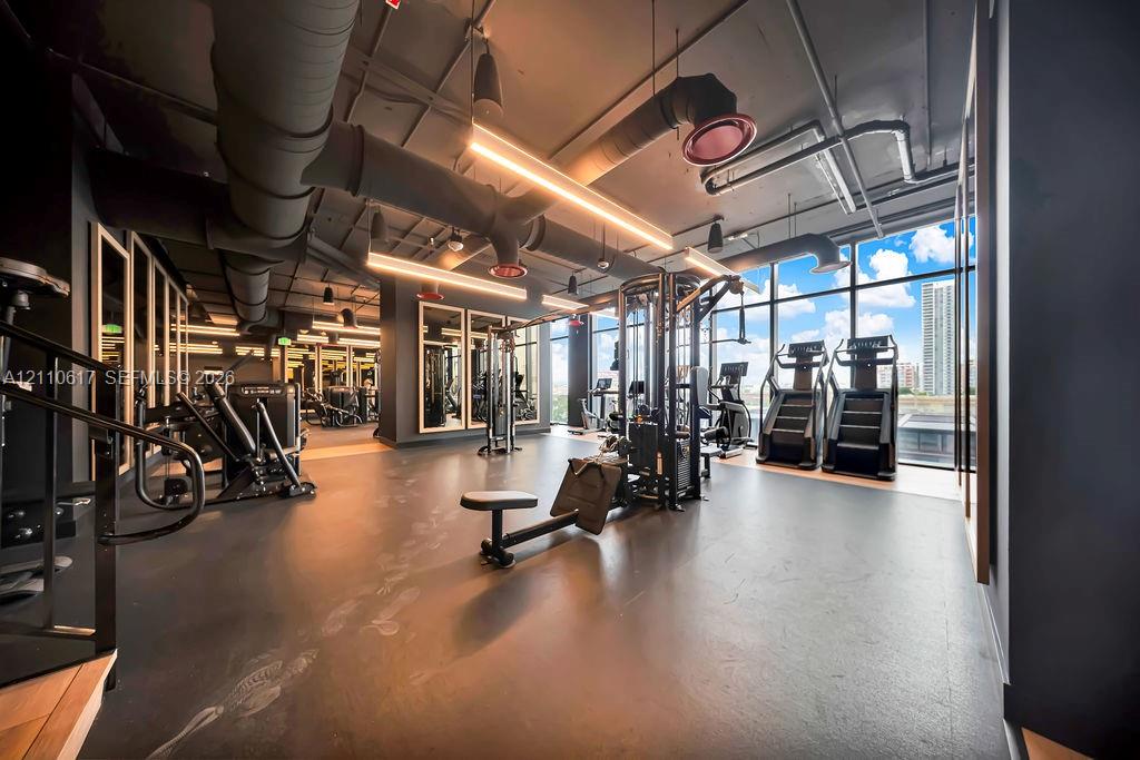 Fitness facility featuring industrial-style exposed ceiling ductwork, rubberized flooring, floor-to-ceiling windows, and integrated linear lighting fixtures