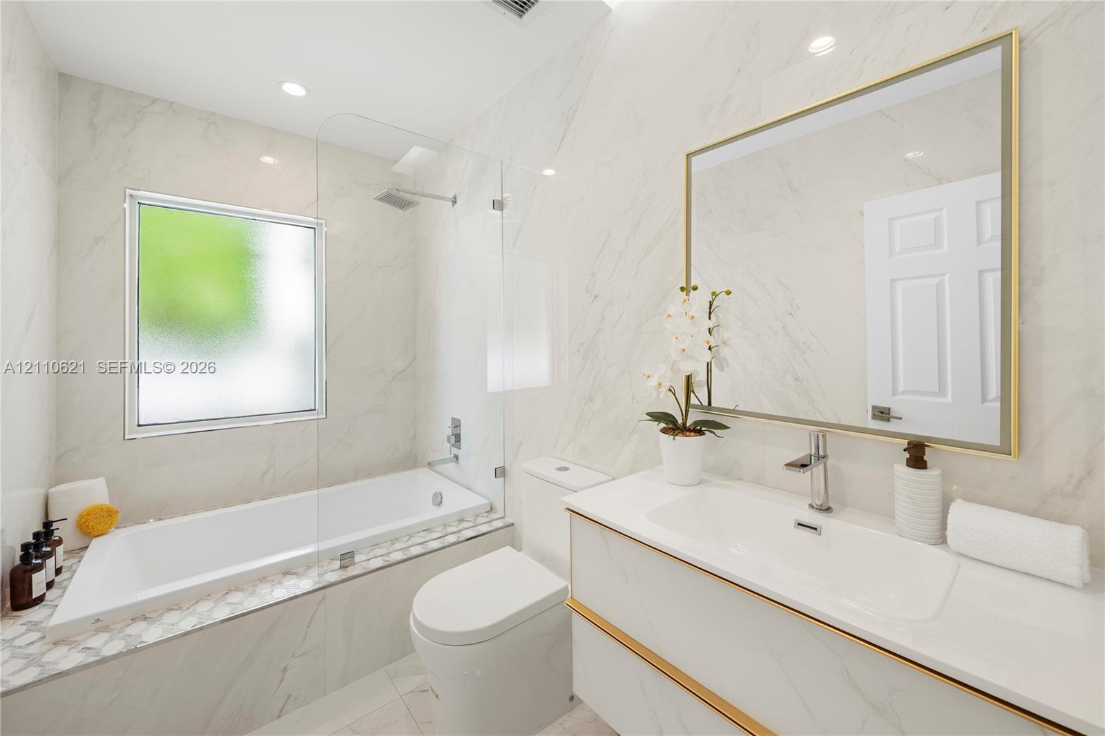 Full-height stone-finish wall tile, white vanity with gold-tone hardware, frameless glass shower enclosure, integrated soaking tub, and gold-framed wall mirror