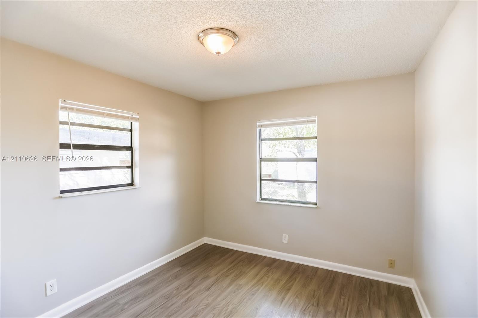 Room featuring wood-finish flooring, neutral wall paint, two windows with horizontal blinds, and a centered flush-mount ceiling light