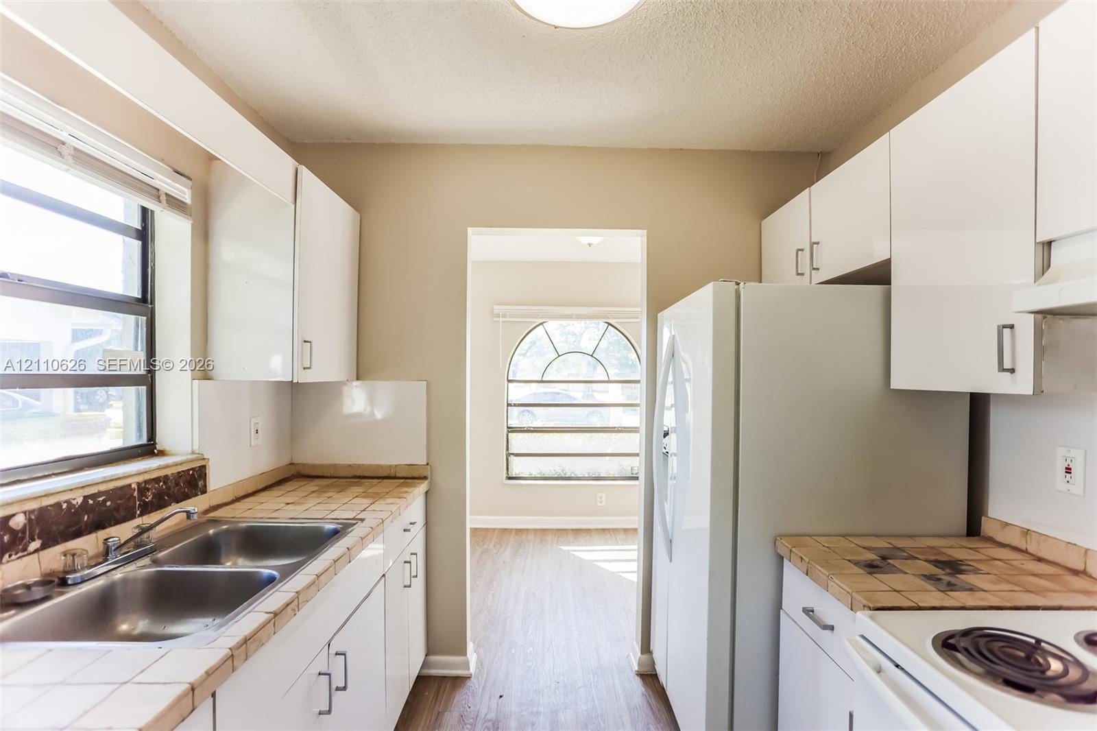 Kitchen featuring white cabinetry, tile countertops, a double-basin stainless steel sink, and an arched window