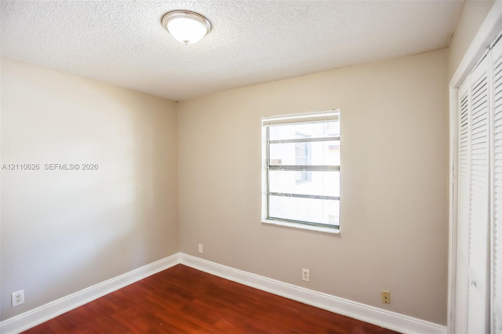 Room featuring wood-finish flooring, neutral wall paint, a single-pane window, flush-mount ceiling lighting, and louvered bifold closet doors