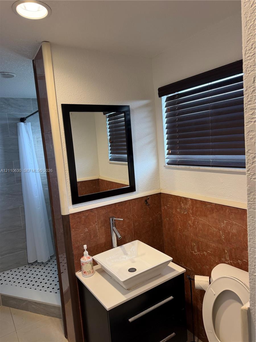 Modern vanity featuring a square vessel sink with a chrome faucet, topped with a countertop, complemented by a framed mirror, window blinds, and tile wainscoting