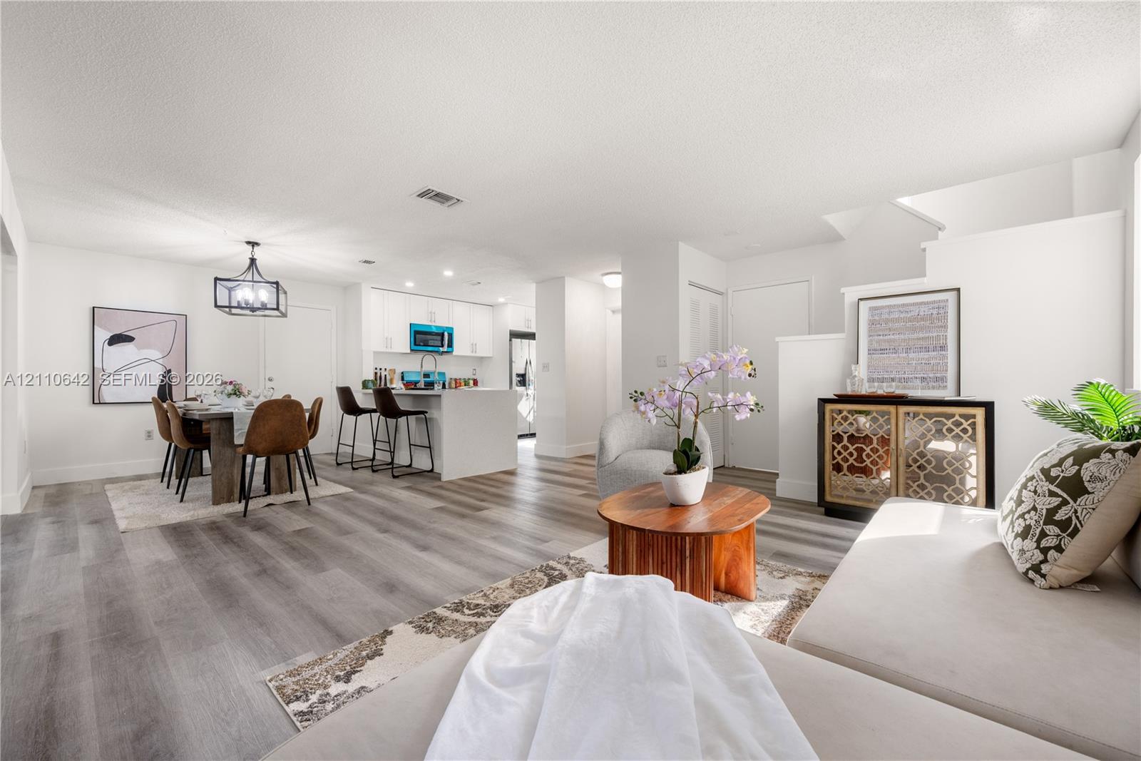 Open-concept interior featuring wood-finish flooring, white cabinetry, a kitchen island with stone-surface countertop, and a contemporary geometric hanging light fixture Open-concept interior featuring wood-finish flooring, white cabinetry, a kitchen island with stone-surface countertop, and a contemporary geometric hanging light fixture