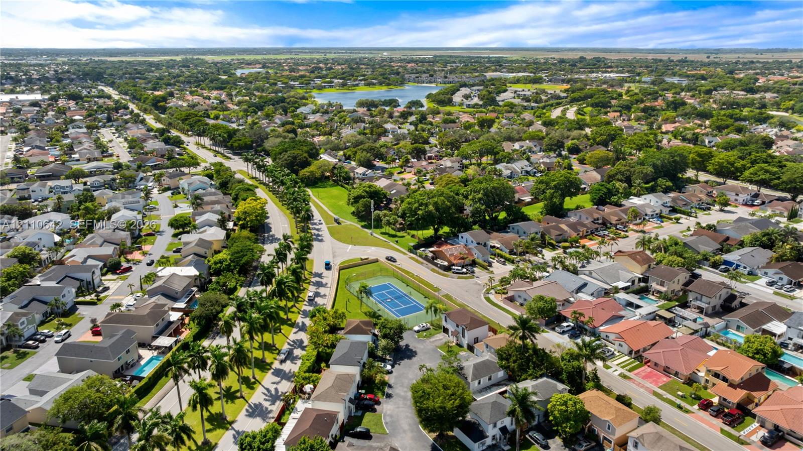 Aerial perspective of a residential community featuring a fenced tennis court, mature landscaping, and proximity to a central lake