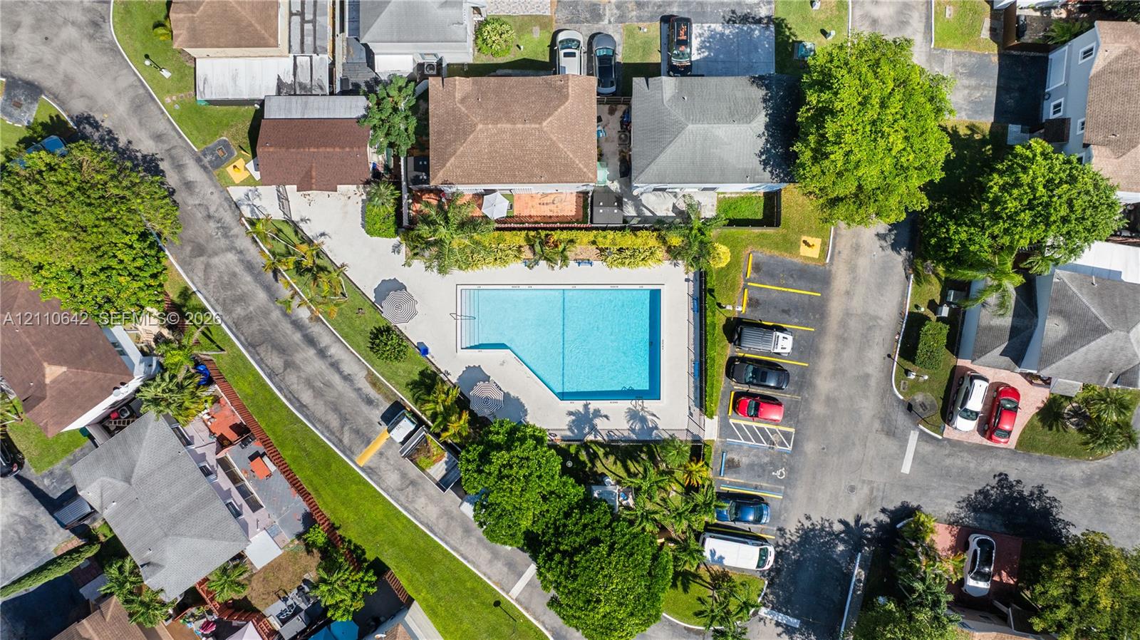 Aerial perspective of a community swimming pool featuring a paved pool deck, perimeter fencing, and surrounding mature landscaping