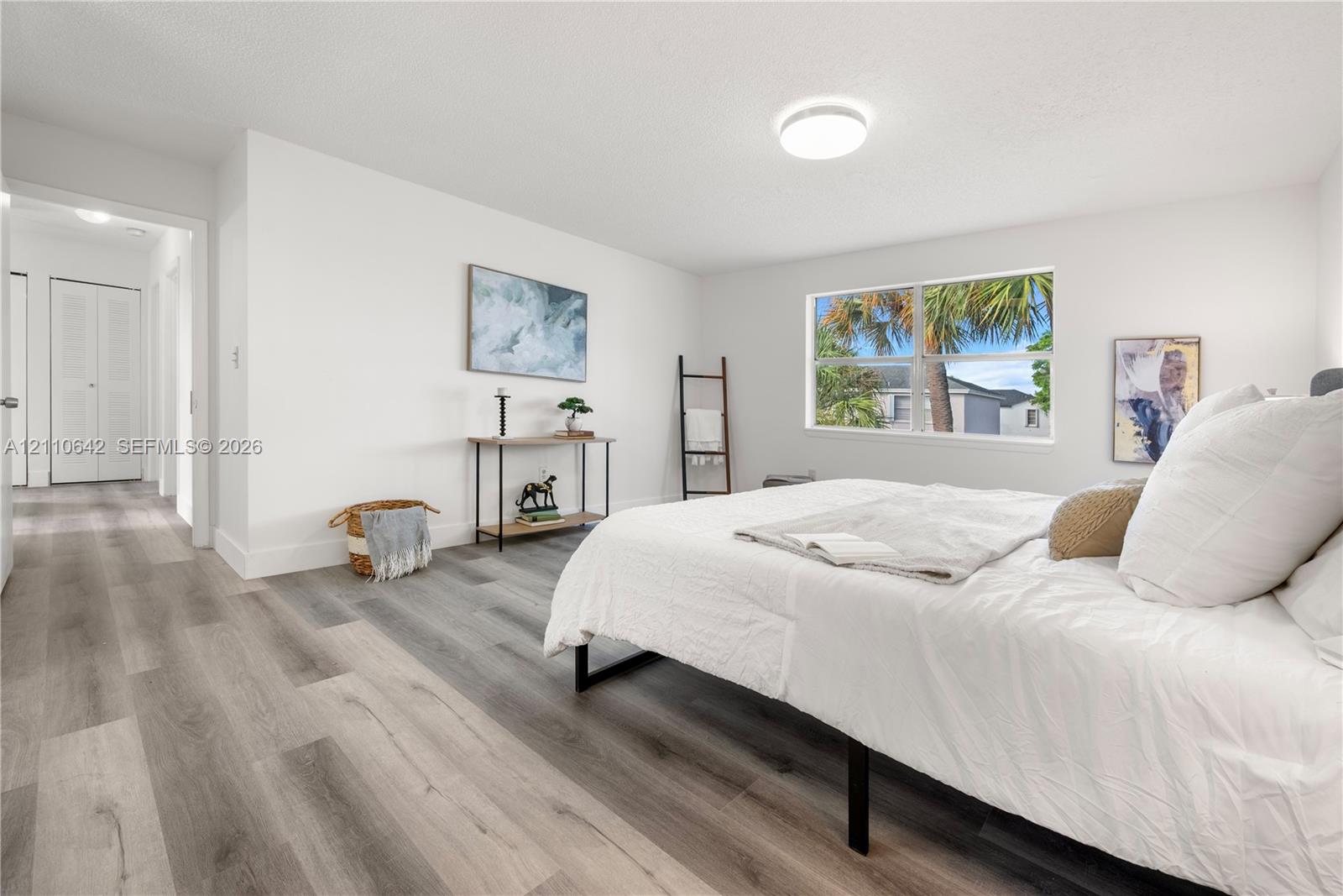 Neutral-toned wood-finish flooring, crisp white walls, dual-panel closet doors, and large single-hung window featuring outdoor palm tree greenery