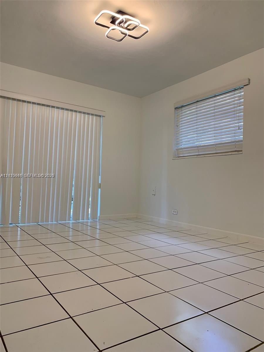 Neutral-toned room featuring large-format ceramic tile flooring, modern geometric LED flush-mount lighting, sliding glass patio doors, and horizontal window blinds Neutral-toned room featuring large-format ceramic tile flooring, modern geometric LED flush-mount lighting, sliding glass patio doors, and horizontal window blinds