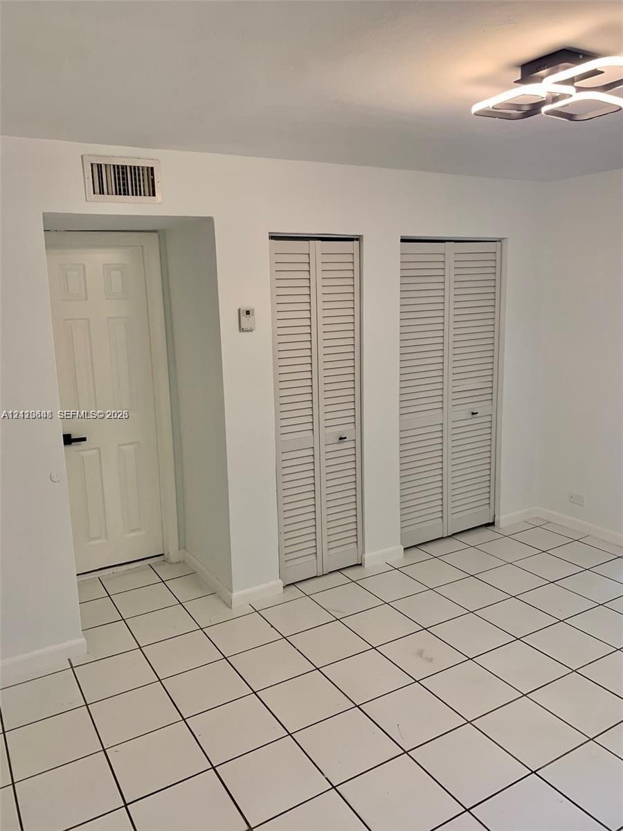 Neutral-toned room featuring large-format square tile flooring, two sets of louvered bifold closet doors, a single-panel interior door with dark hardware, and a modern geometric flush-mount ceiling light Neutral-toned room featuring large-format square tile flooring, two sets of louvered bifold closet doors, a single-panel interior door with dark hardware, and a modern geometric flush-mount ceiling light