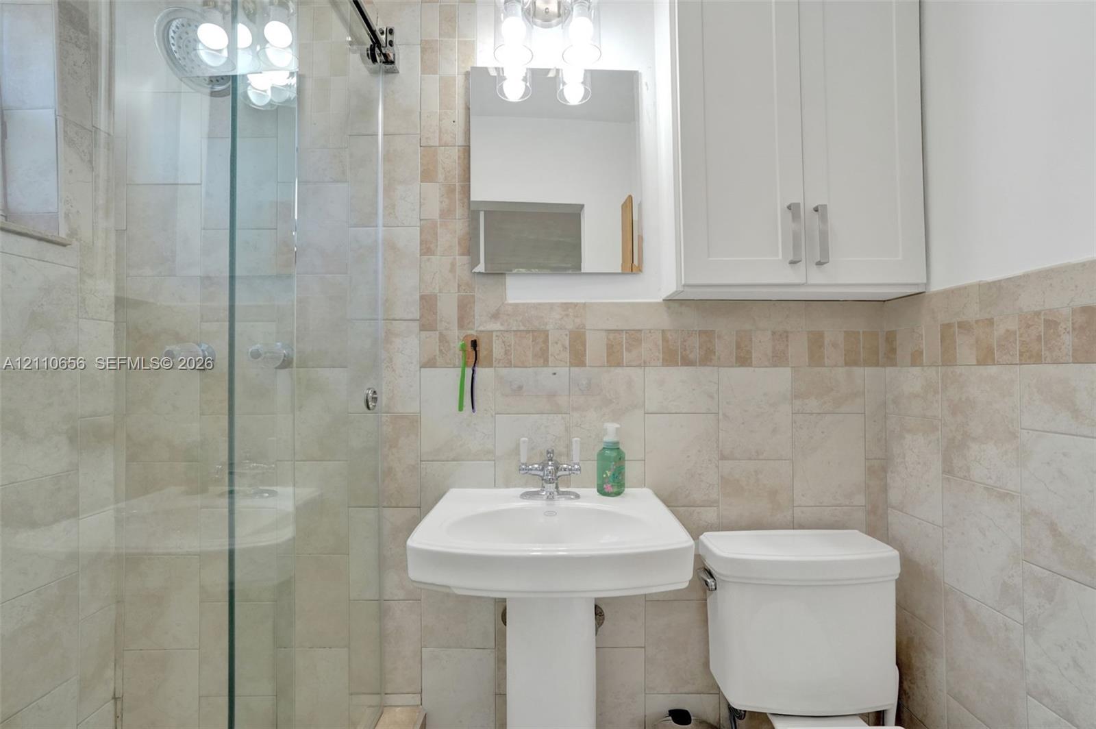 Glass-enclosed shower with stone tile surround, pedestal sink with polished chrome hardware, built-in white wall cabinetry, and coordinating stone wall tiling