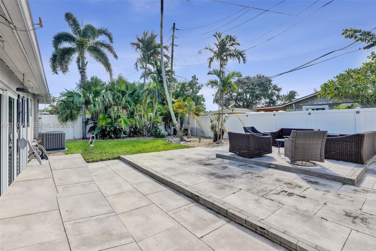 Backyard featuring a split-level concrete paver patio, a grass lawn, mature palm trees, and perimeter vinyl fencing