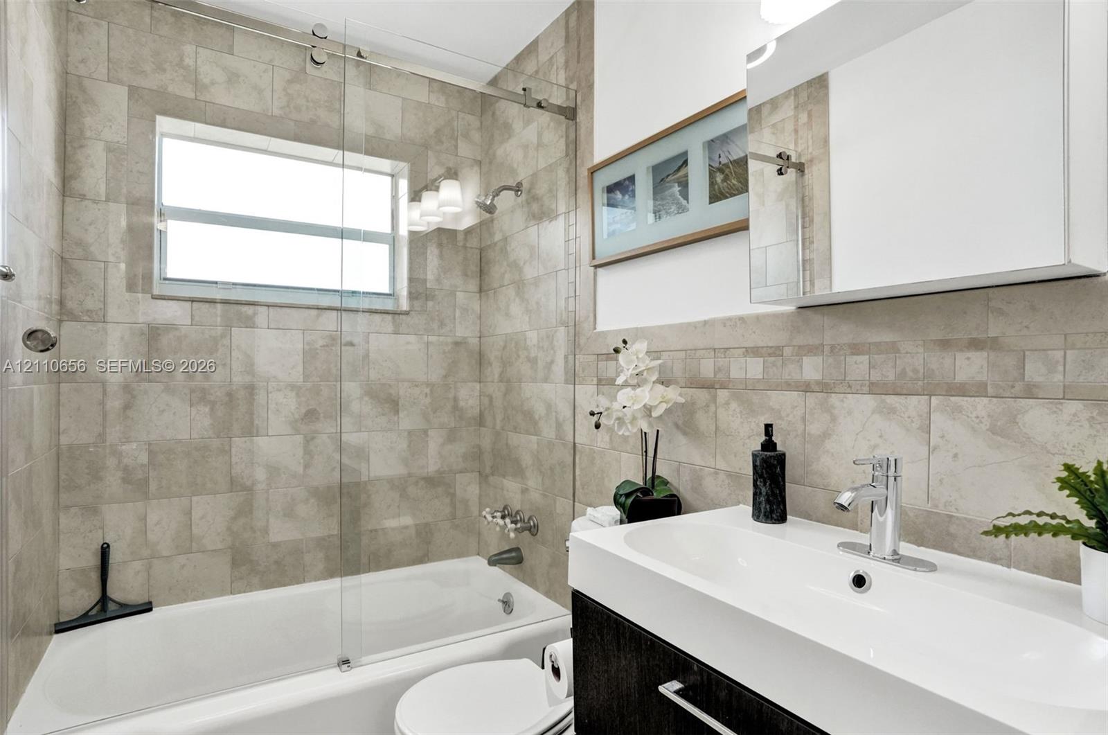 Bathroom featuring a glass sliding shower door, neutral wall tile with a mosaic accent border, white undermount sink with chrome fixtures, and dark wood-finish cabinetry