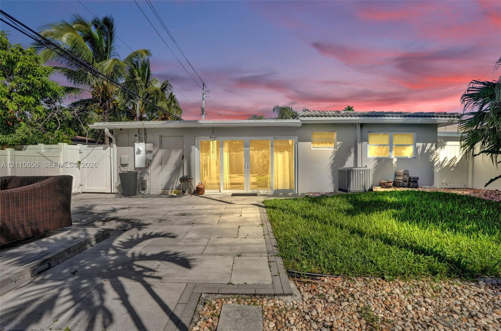Exterior featuring a paved stone patio, multi-panel glass sliding doors, vinyl perimeter fencing, and established palm trees
