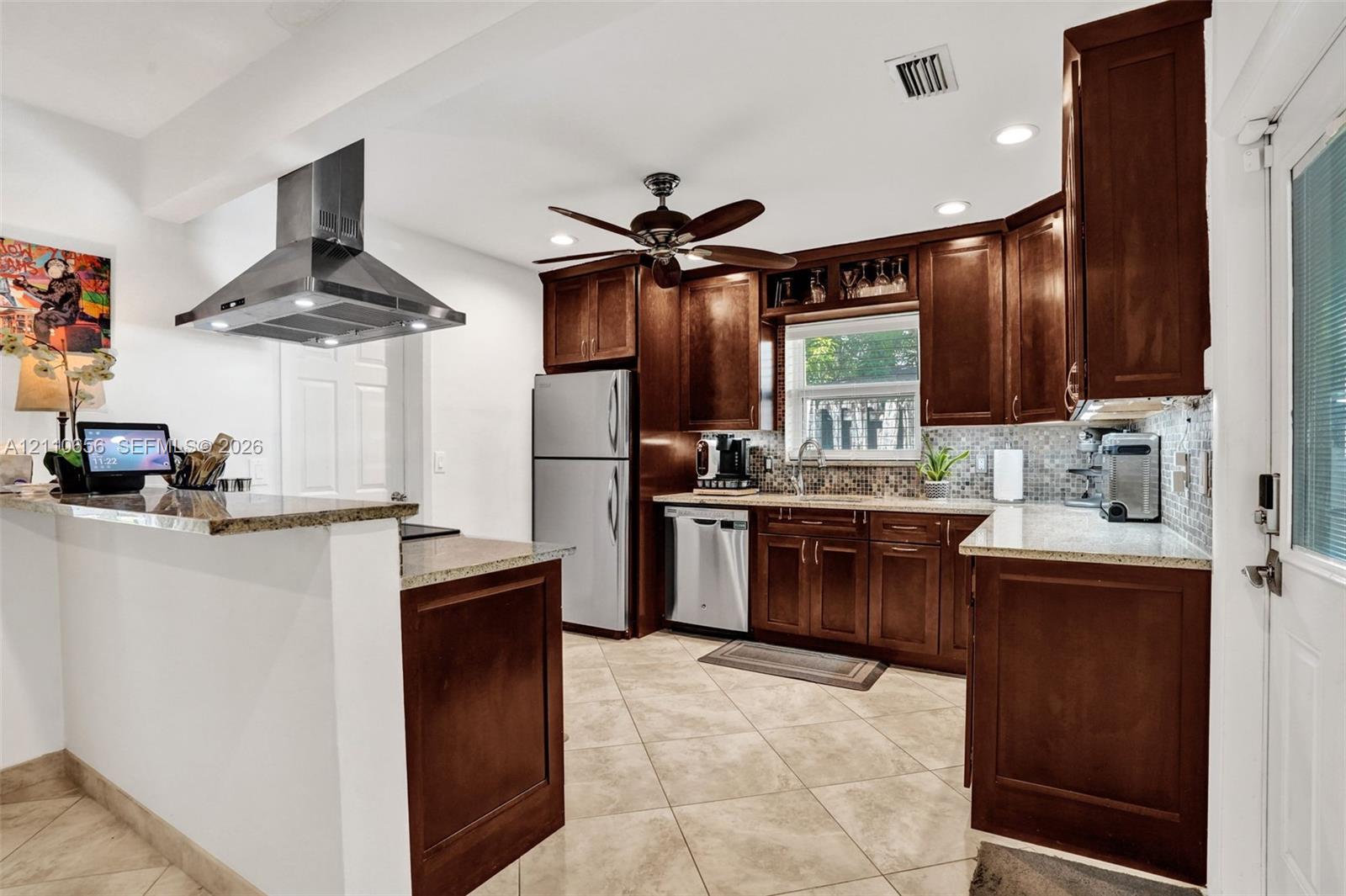 Kitchen featuring dark wood cabinetry, stone countertops, a mosaic tile backsplash, stainless steel appliances, and a ceiling-mounted stainless steel vent hood