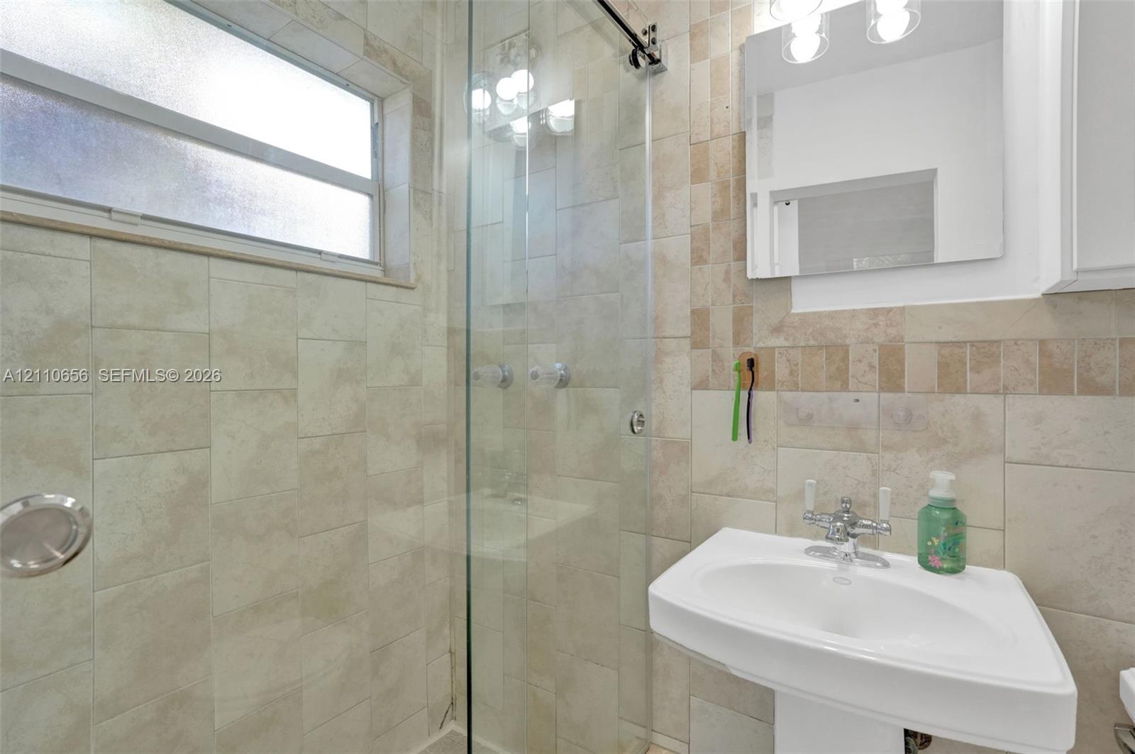 Bathroom featuring a frameless glass shower enclosure, neutral-toned stone tile surround, pedestal sink with chrome hardware, and horizontal sliding window