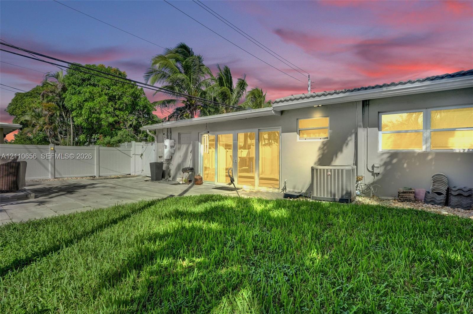 Rear exterior featuring a manicured lawn, paved patio area, white vinyl privacy fencing, sliding glass doors, and mature palm trees