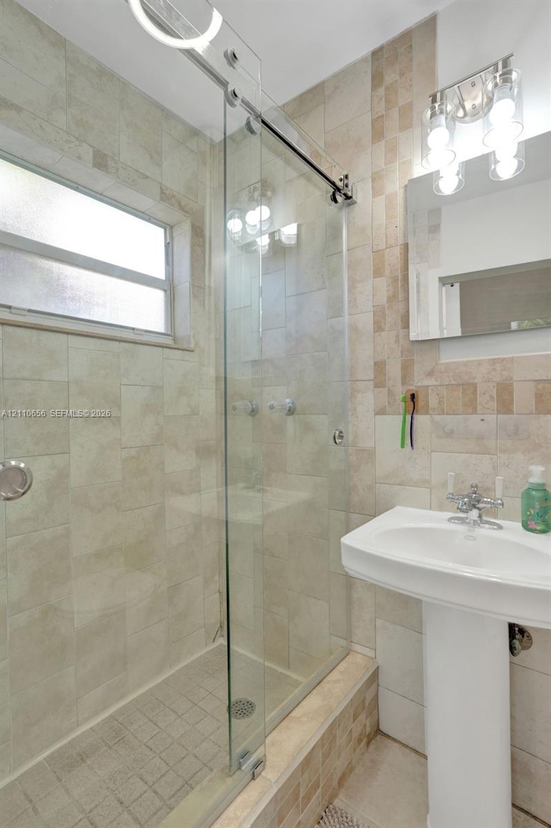 Glass-enclosed shower with a sliding door, pedestal sink, wall-mounted rectangular mirror, and neutral-toned wall tile