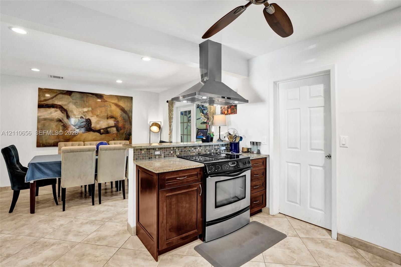 Kitchen featuring dark wood cabinetry, stone countertops, mosaic tile backsplash, stainless steel range with overhead vent hood, and tile flooring