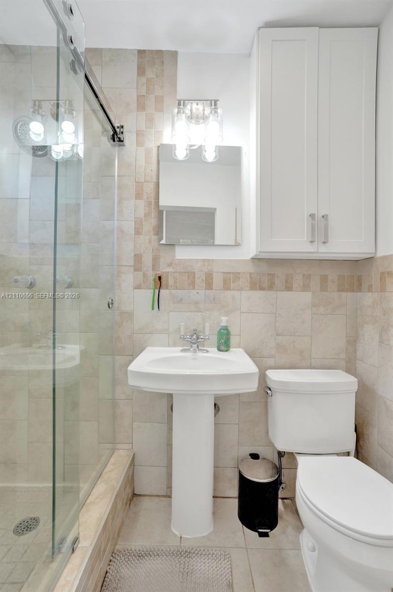 Bathroom featuring neutral stone-finish wall tile with mosaic accents, a glass-enclosed shower, a white pedestal sink, and wall-mounted white cabinetry