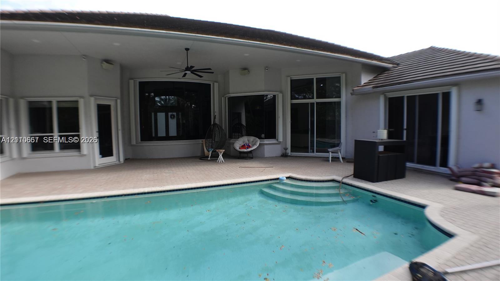 Covered patio with integrated ceiling fan, in-ground pool with stone paver decking, and mature tropical landscaping