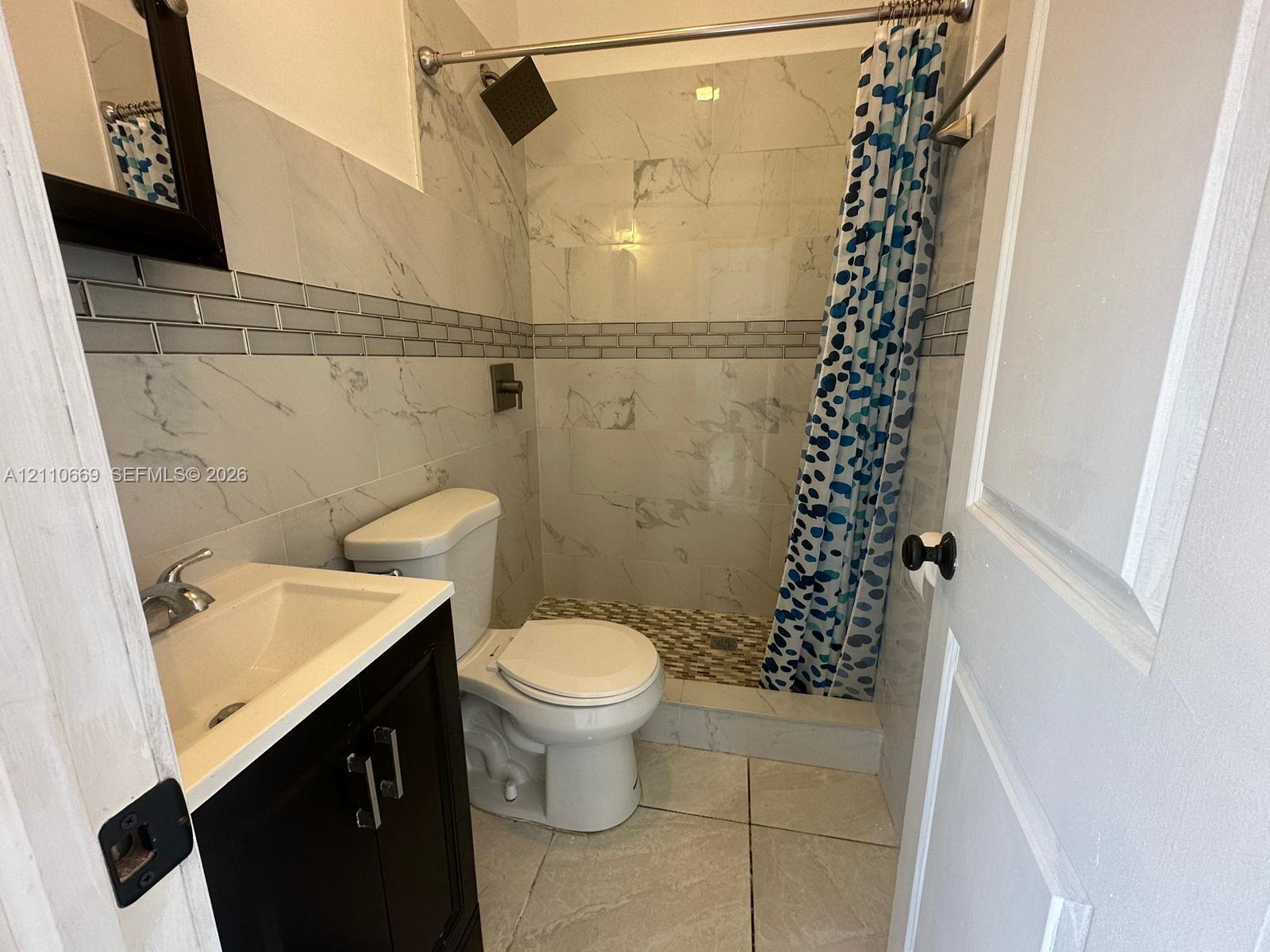 Bathroom featuring a dark-finish vanity with a chrome faucet, large-format wall tile with decorative glass mosaic inlay, a walk-in shower with stone-style flooring, and a fixed rain-style showerhead