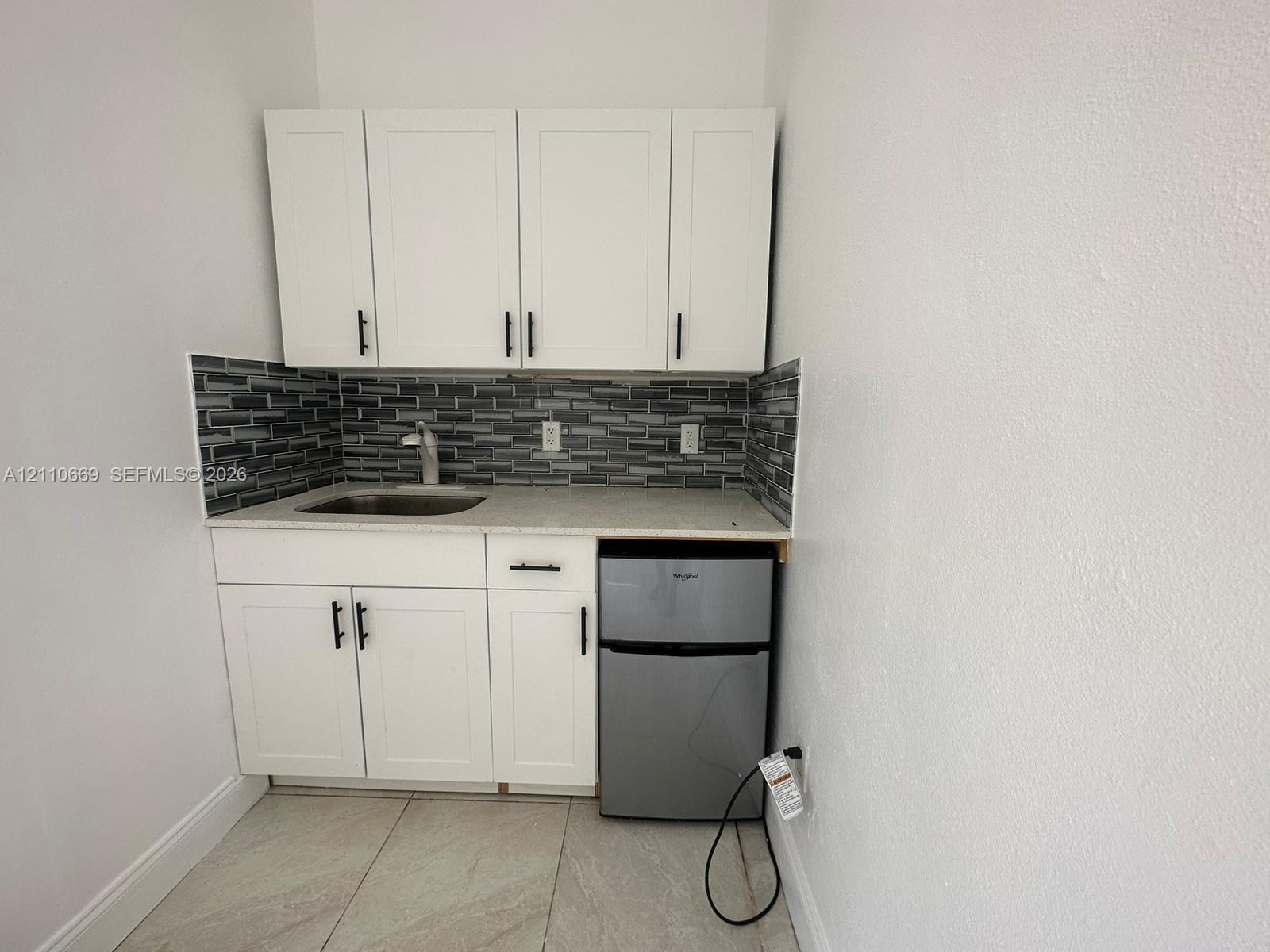 Kitchenette featuring white cabinetry with matte black hardware, stone-finish countertop, stainless steel sink with high-arc faucet, linear mosaic tile backsplash, and built-in under-counter refrigerator