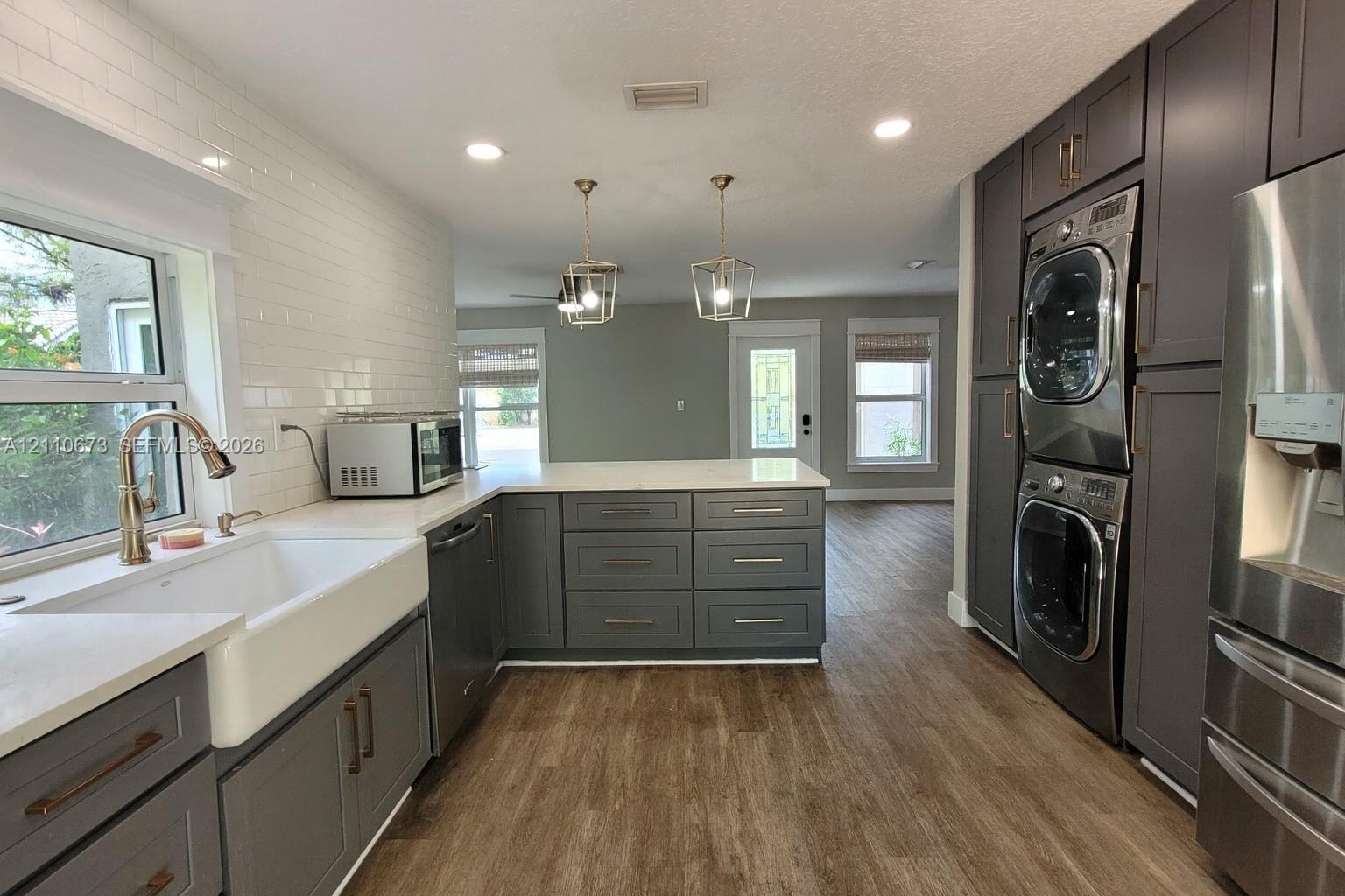 Kitchen featuring dark gray cabinetry, white subway tile backsplash, farmhouse-style sink, quartz countertops, and built-in stacked laundry appliances Kitchen featuring dark gray cabinetry, white subway tile backsplash, farmhouse-style sink, quartz countertops, and built-in stacked laundry appliances
