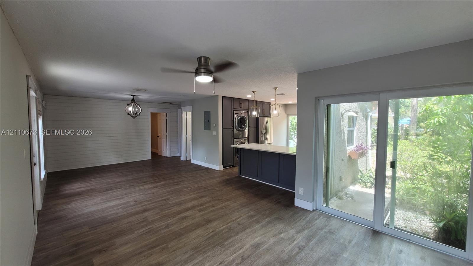 Open-concept interior featuring wood-finish flooring, a shiplap accent wall, kitchen island with quartz-style countertop, ceiling fan with integrated lighting, and sliding glass doors Open-concept interior featuring wood-finish flooring, a shiplap accent wall, kitchen island with quartz-style countertop, ceiling fan with integrated lighting, and sliding glass doors