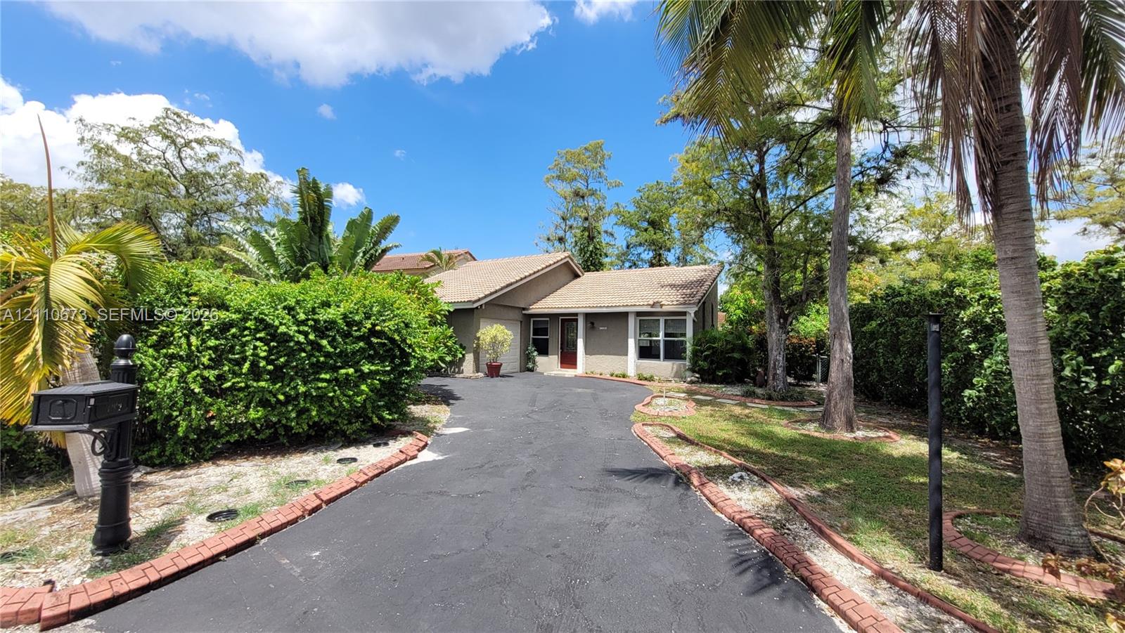 Asphalt driveway framed by a red brick border, established landscaping, mature palm trees, and exterior siding