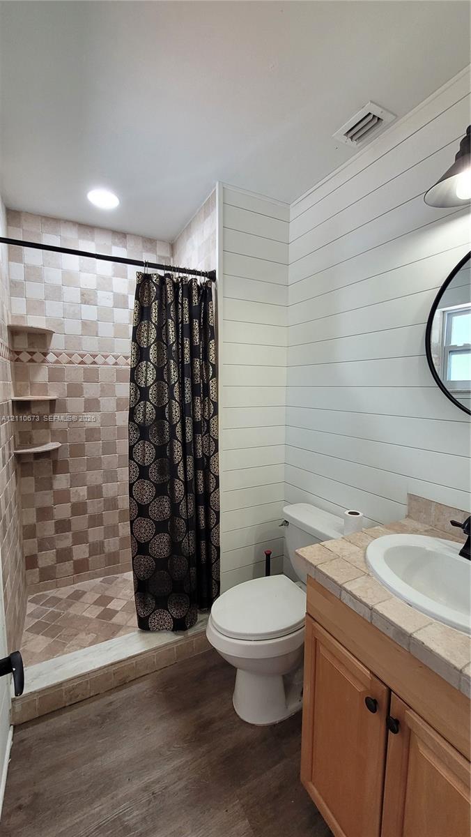 Bathroom featuring white shiplap walls, wood-finish flooring, a tiled vanity countertop with an integrated basin, and a walk-in shower with built-in shelving and multi-toned wall tile Bathroom featuring white shiplap walls, wood-finish flooring, a tiled vanity countertop with an integrated basin, and a walk-in shower with built-in shelving and multi-toned wall tile